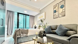 A modern hotel room with a large bed, a grey sofa, a small side table, and a view of the city through large windows. - Cosmos Living Delightful Studio (Dubai)