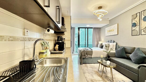 A modern hotel room with a kitchenette, a sofa, a coffee table, and a large window offering a city view. - Cosmos Living Delightful Studio (Dubai)