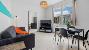 Ein modernes Wohnzimmer mit Sofa, Fernseher, Esstisch, Stühlen und einem großen Fenster mit Vorhängen.