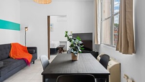Ein modernes Wohnzimmer mit einem dunklen Sofa, einem Tisch mit Stühlen, einer Hängeleuchte und einem großen Fenster mit Vorhängen.
