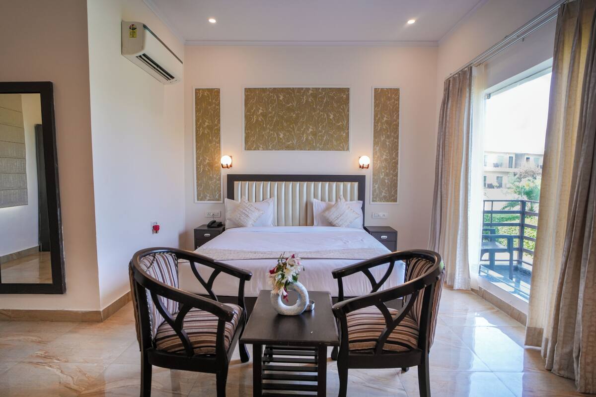 Habitación de hotel con una cama grande, dos mesitas de noche, una pequeña mesa con sillas, un equipo de aire acondicionado y vistas al exterior.