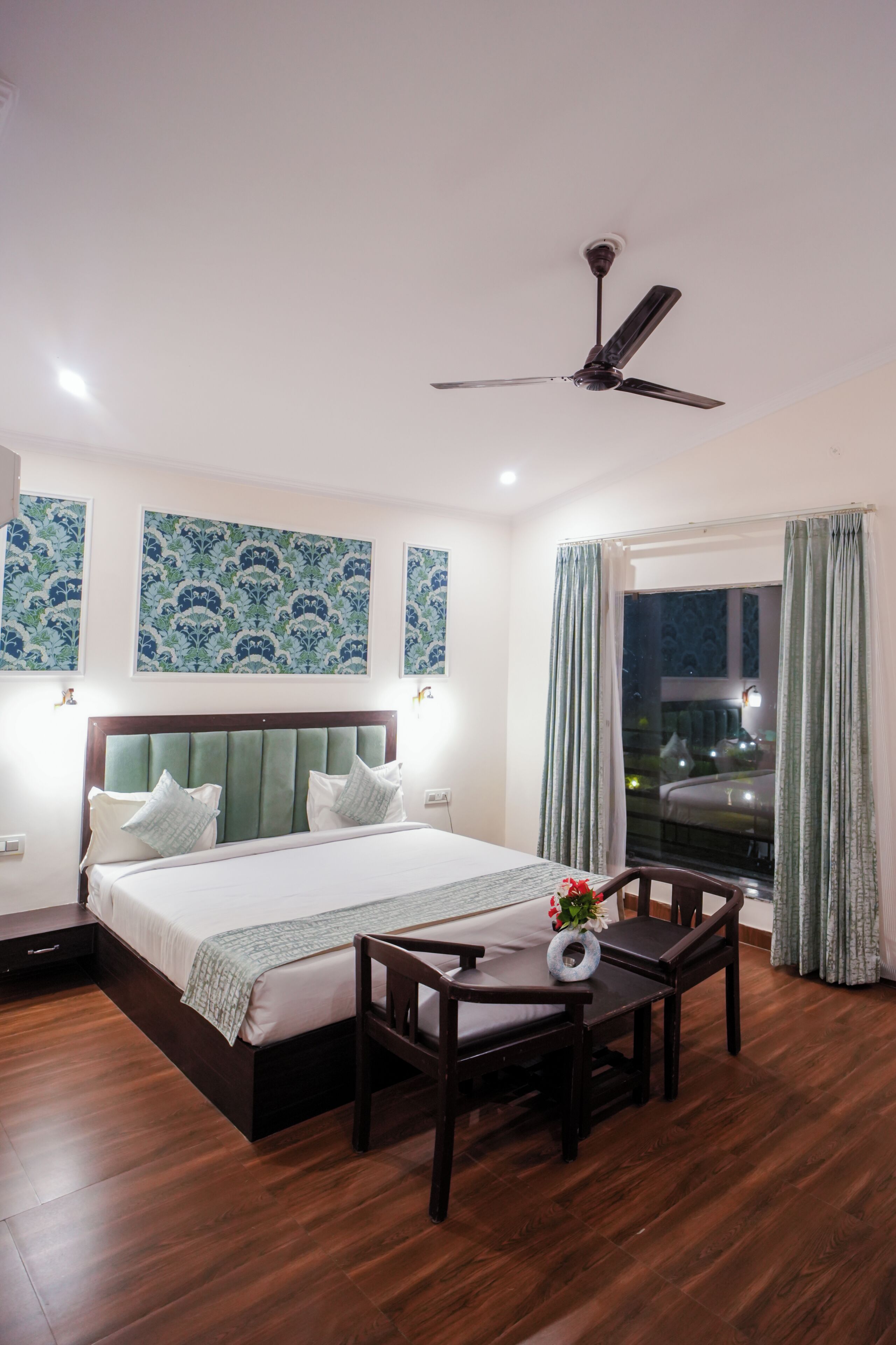 Una habitación de hotel con cama, una mesa y sillas, ventilador de techo y ventana con cortinas.