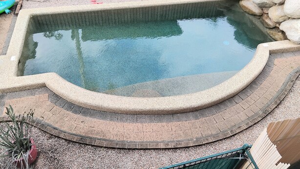 A small, rectangular swimming pool with a stone edge, surrounded by a pebble bed and a wooden fence.