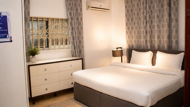 Une chambre à coucher comprenant un lit, une lampe de chevet, un appareil de climatisation, une fenêtre avec des rideaux et une commode.