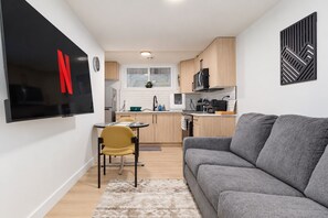 Un espacio habitable compacto con kitchenette, un sofá gris, una silla amarilla y un televisor de pantalla plana montado en la pared.