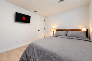 A bedroom with a bed, a wall-mounted TV, a door, and a nightstand with a lamp.