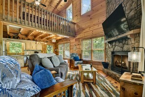 A wooden cabin interior with a living room, kitchen, and stone fireplace.