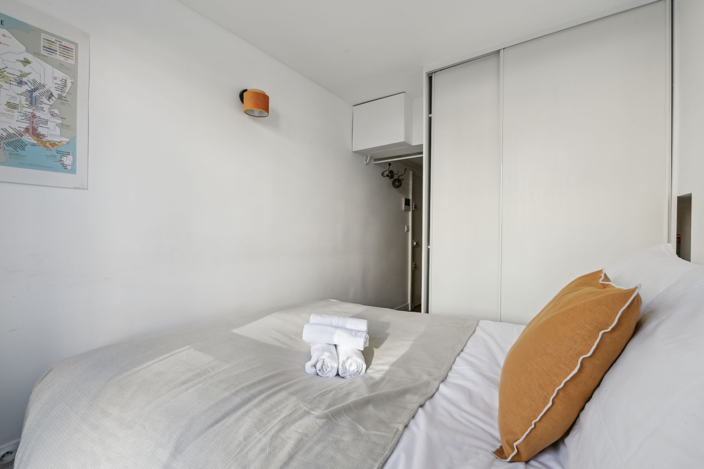 Un lit bien fait, avec du linge de lit blanc et un oreiller marron, une carte accrochée au mur et une porte de salle de bain.