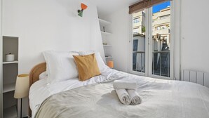 A neatly made bed with white bedding and a folded towel, a bedside lamp, and a window with a view of buildings.