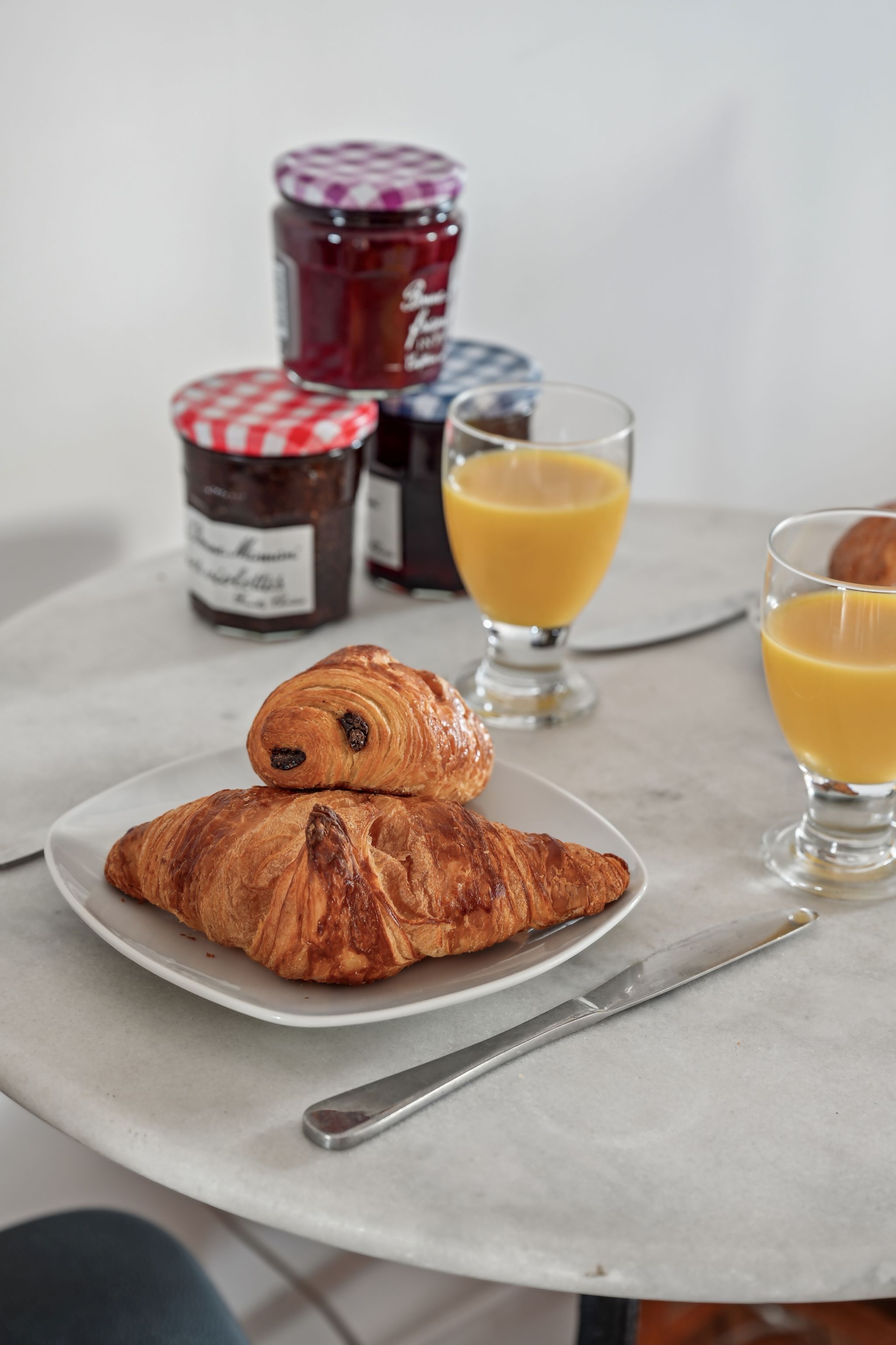 A breakfast setting with a croissant, two glasses of orange juice, and jars of jam on a table.