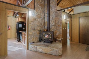 A stone fireplace with a wood-burning stove, wooden beams, and a kitchen area with appliances. - 7 Bobcat - Hot Tub, SHARC, Close to Village, A/C (Sunriver)