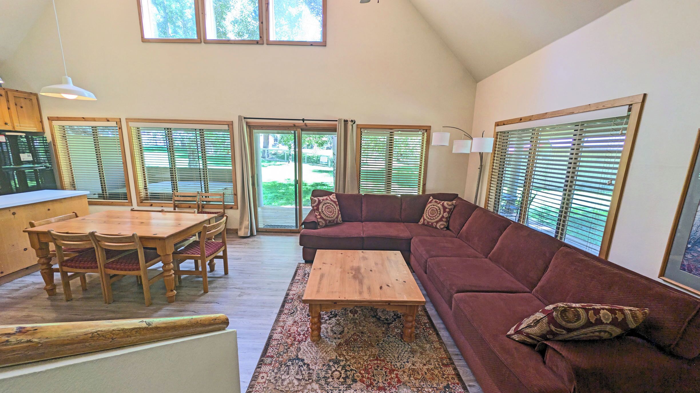A spacious living area with a wooden dining table, a red sofa, and a coffee table. There are multiple windows with blinds, a ceiling fan, and a kitchen area visible in the background.