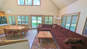 A spacious living area with a wooden dining table, a red sofa, and a coffee table. There are multiple windows with blinds, a ceiling fan, and a kitchen area visible in the background. - Circle Four Condo 14 - Hot Tub, SHARC Passes, Community Pool (Sunriver)