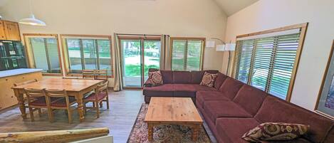 A spacious living area with a wooden dining table, a red sofa, and a coffee table. There are multiple windows with blinds, a ceiling fan, and a kitchen area visible in the background.