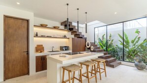 A modern kitchen with a central island, wooden stools, and a staircase leading to a balcony with plants. - La Finca Bingin (Pecatu)