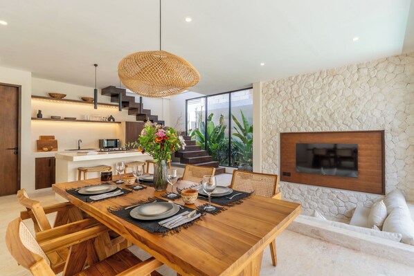 A modern kitchen with a dining area, featuring a wooden table set for six, a large woven pendant light, and a stone accent wall. - La Finca Bingin (Pecatu)