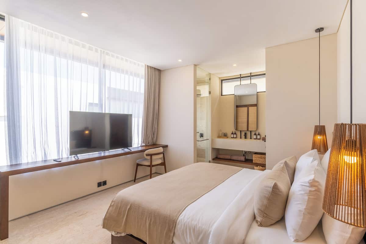 A modern hotel room with a large bed, a desk with a chair, a large window with curtains, a bathroom with a mirror and sink, and a hanging light fixture.