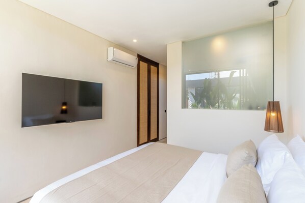 A modern bedroom with a large bed, a mounted TV, and a window with a view of greenery. - La Finca Bingin (Pecatu)