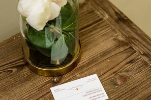A glass dome with a white flower and green leaves, placed on a wooden surface with a business card beside it. - Tresca Charme - Suite 11 Elegant Stay in the Heart of Bari (Bari)