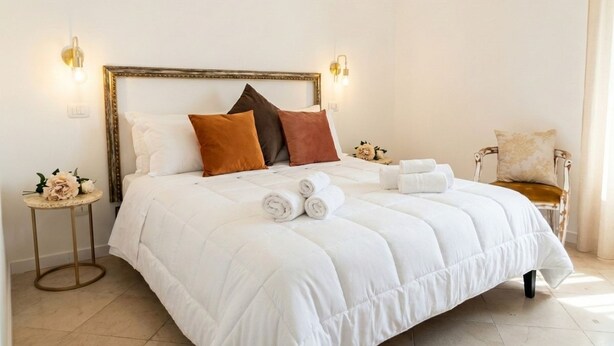 A neatly made bed with a white duvet, brown and white pillows, and rolled towels on a white headboard.