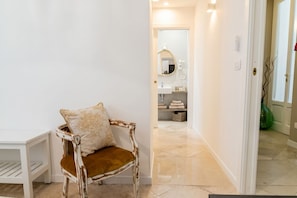 A hallway with a white door, a chair with a cushion, and a bathroom visible in the background.