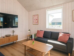 A living room with a flat-screen TV, a wooden cabinet, a sofa with cushions, a small table with a vase, and a window with a view of a house. - 5 Star Holiday Home in Jerup (Jerup)