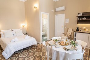 A hotel room with a bed, a dining table set for two, a kitchen area, and a bathroom visible through a glass door. - Tresca Charme - Suite 22-Suite with Private Terrace (Bari)