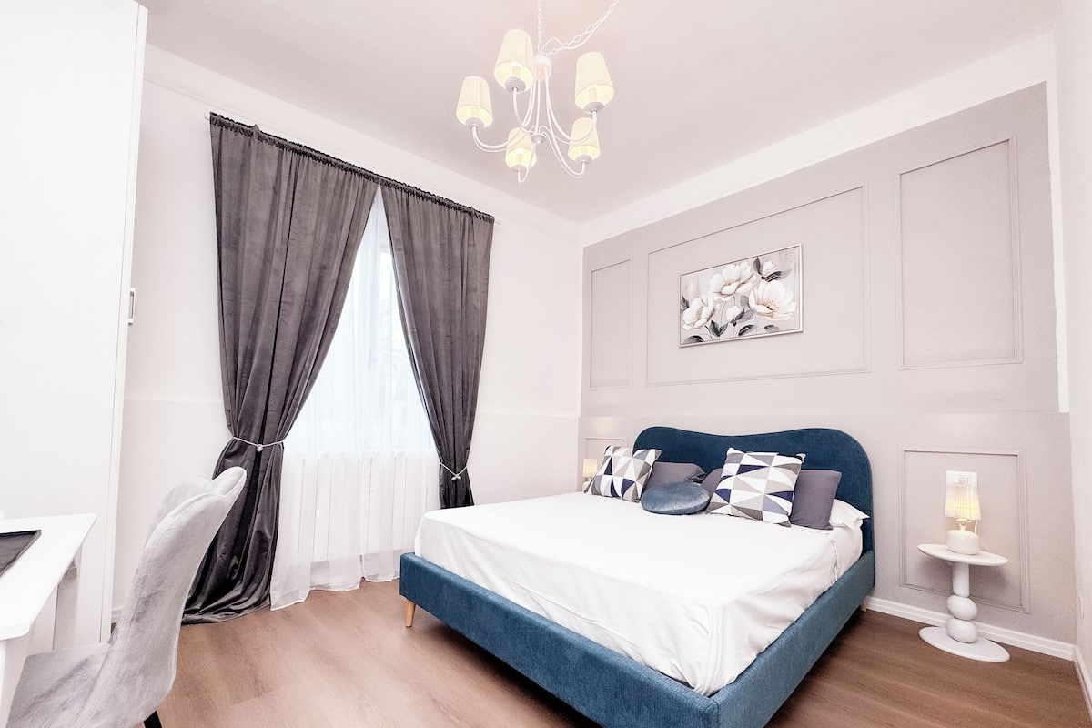 A bedroom with a bed, a desk, a chair, a chandelier, and a window with curtains.
