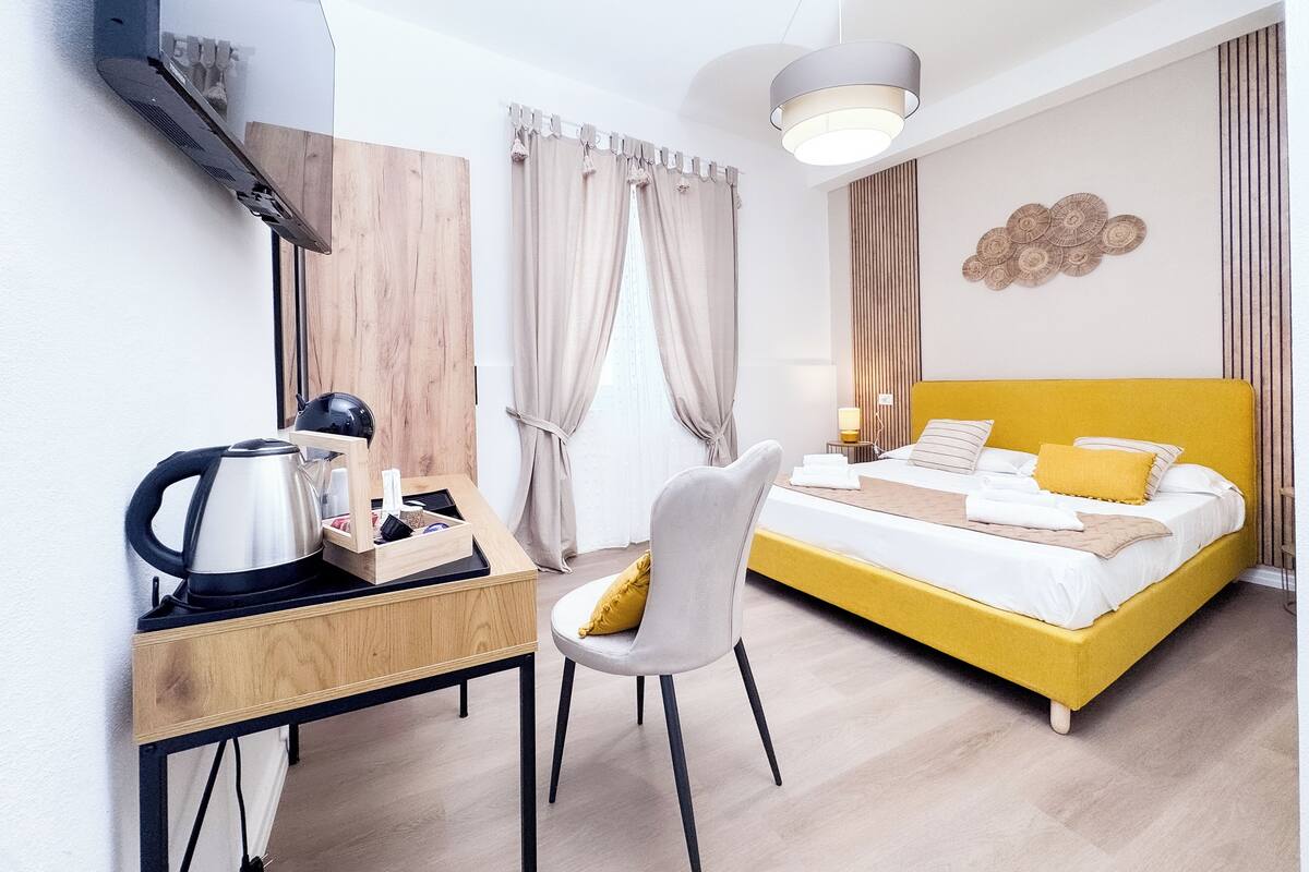 A hotel room with a bed, a desk, a chair, a kettle, and a lamp.