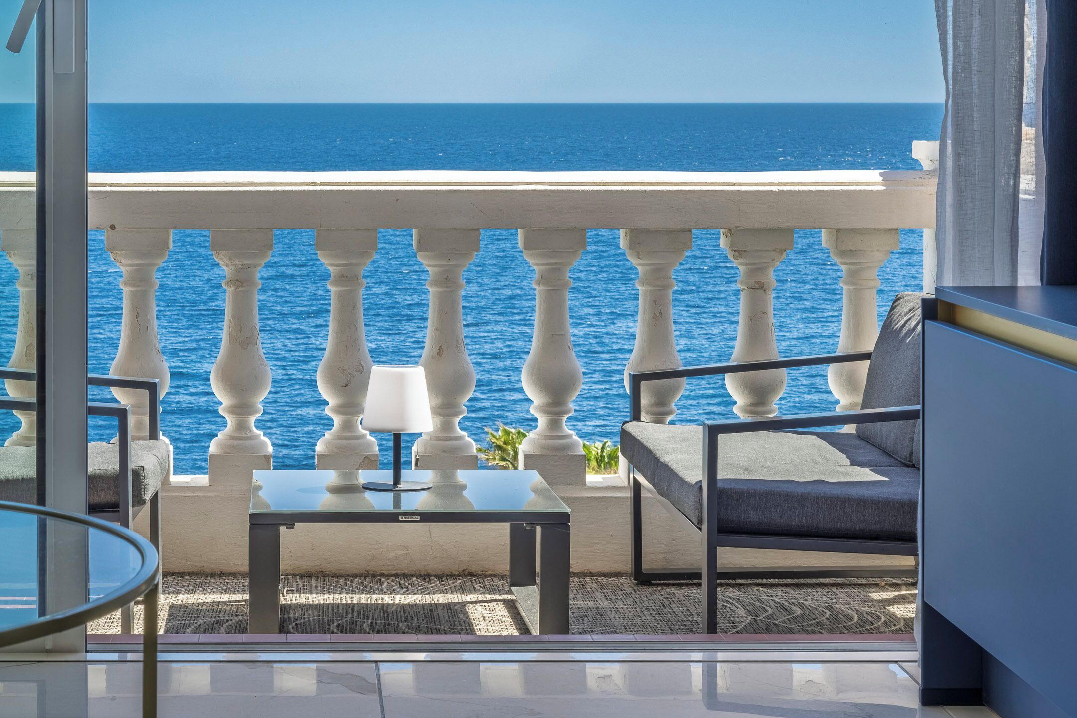 A balcony with a view of the sea, a table with a lamp, and a sofa.