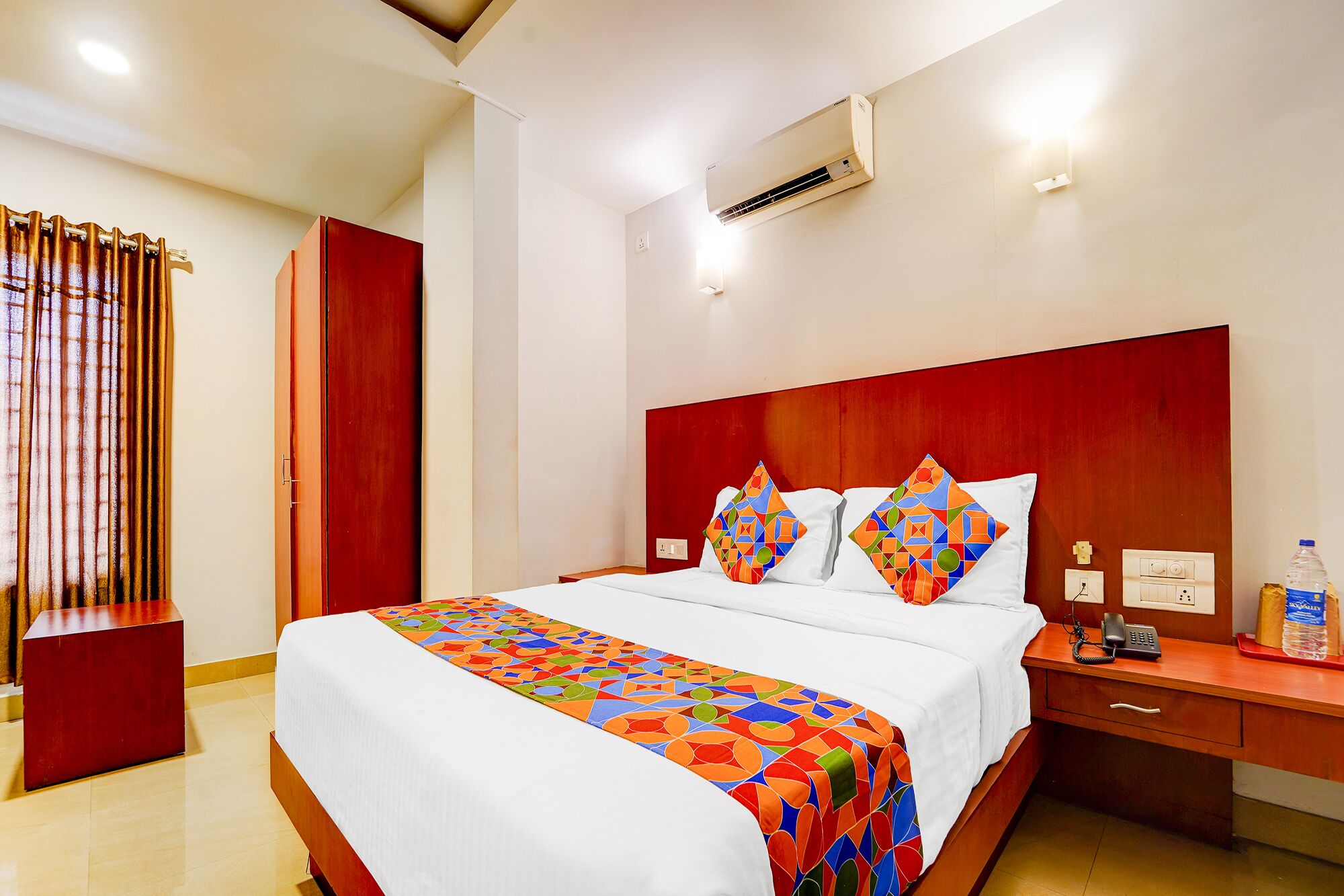 Une chambre d’hôtel avec un grand lit, une table de chevet avec un téléphone et une bouteille, et une armoire rouge.
