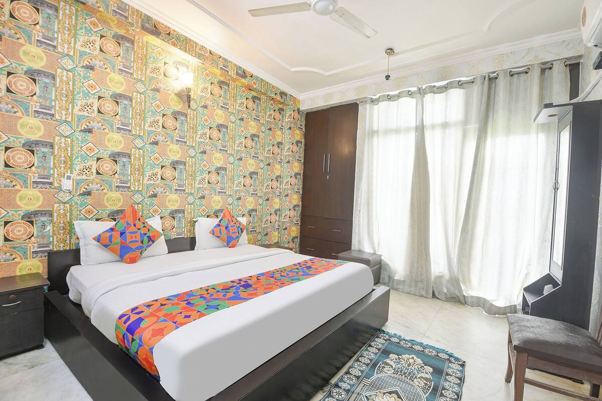 Une chambre à coucher avec un papier peint à motifs, un lit agrémenté de coussins colorés, une armoire en bois et une fenêtre avec des rideaux.