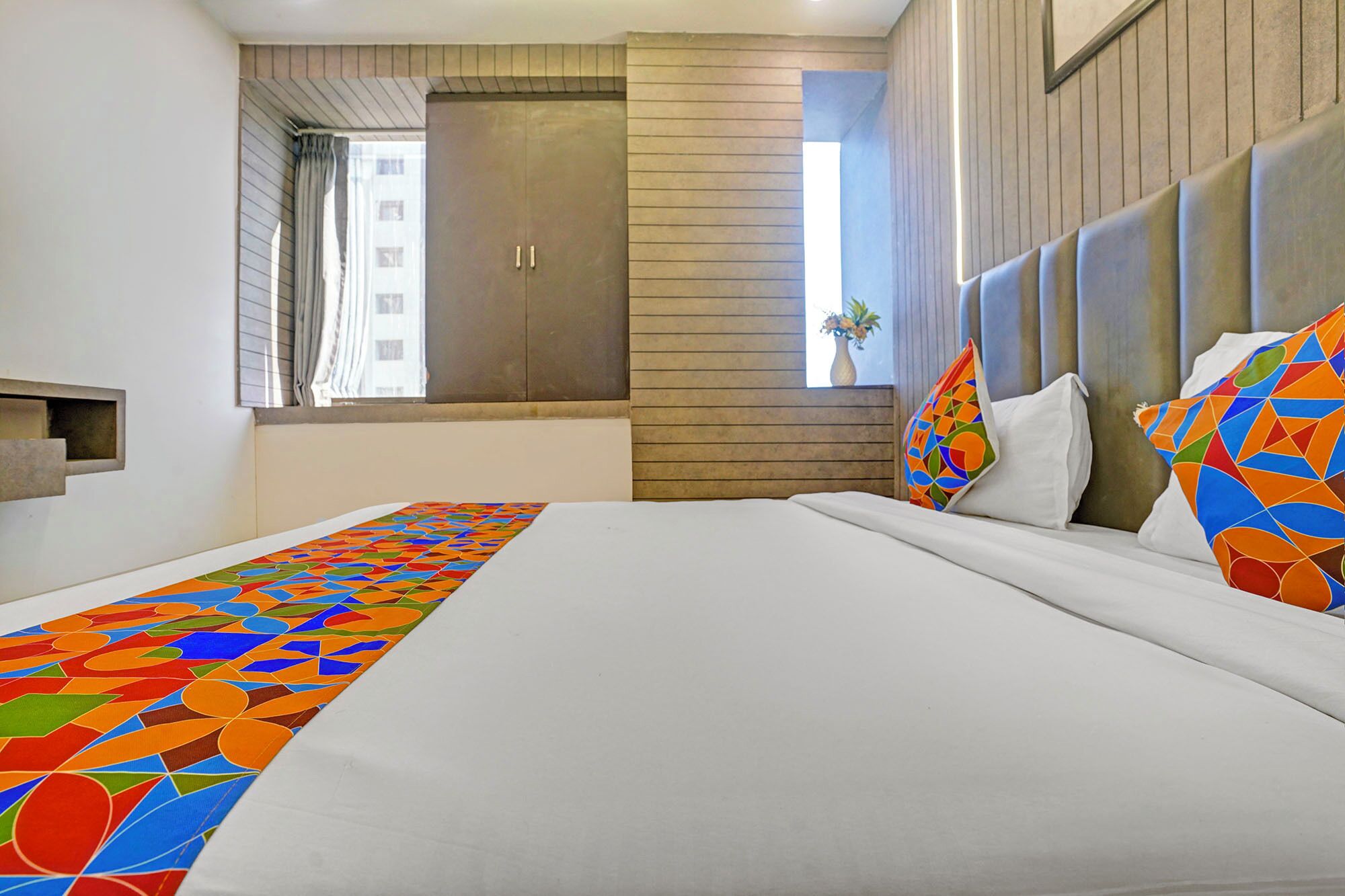 A hotel room with a large bed, a wooden headboard, a window with curtains, and a view of a building.