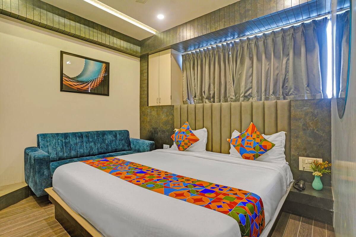 A hotel room with a bed, a blue sofa, a colorful bed runner, a painting on the wall, and a window with curtains.
