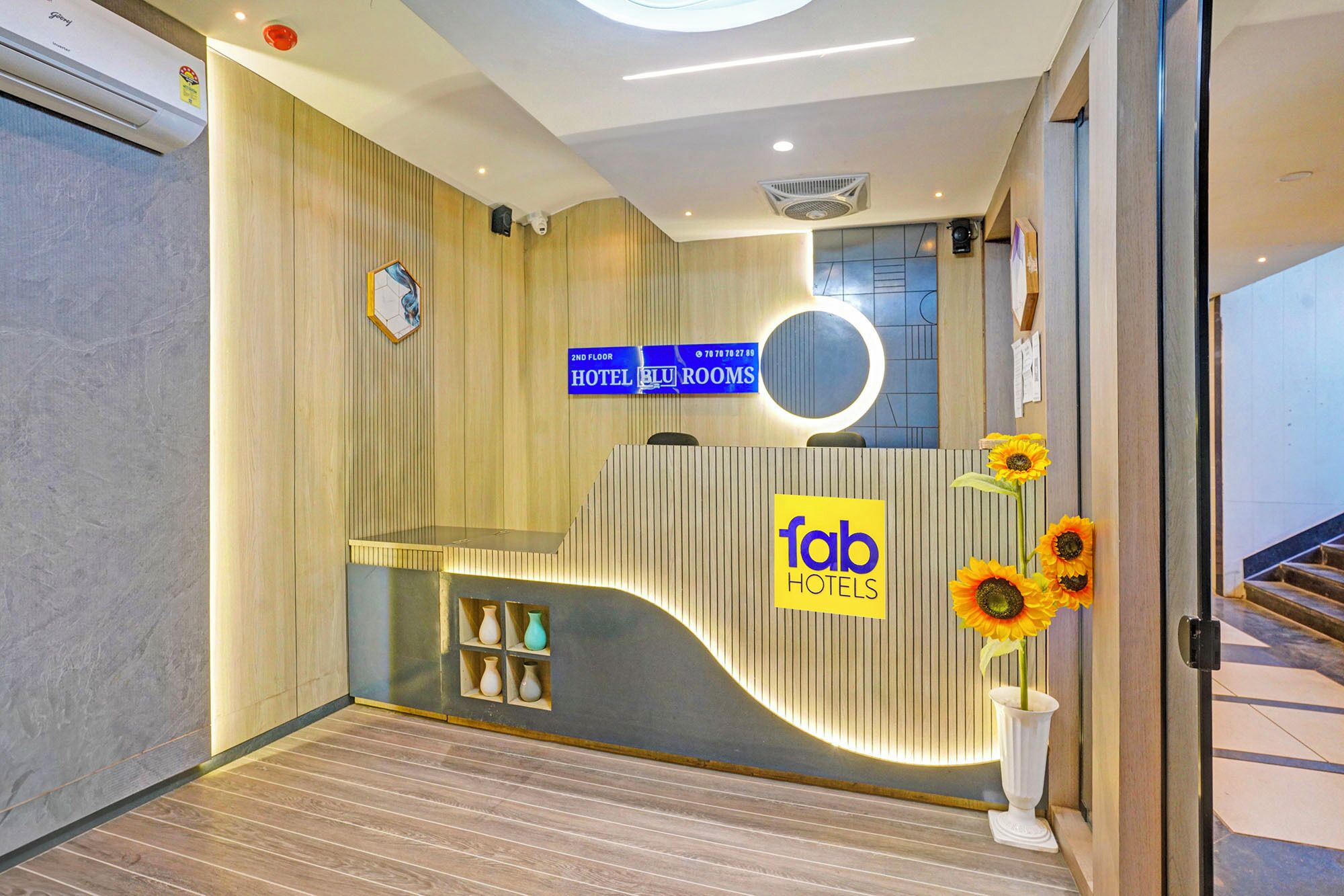 A hotel reception area with a modern design, including a reception desk, a sign for 'fab hotels', and a staircase leading to the second floor.