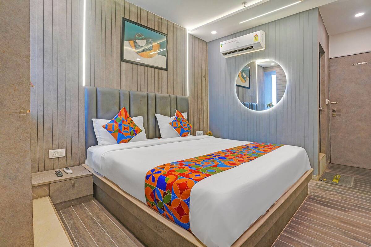 A hotel room with a bed, a nightstand, an air conditioning unit, a wall-mounted artwork, a mirror, and a door.