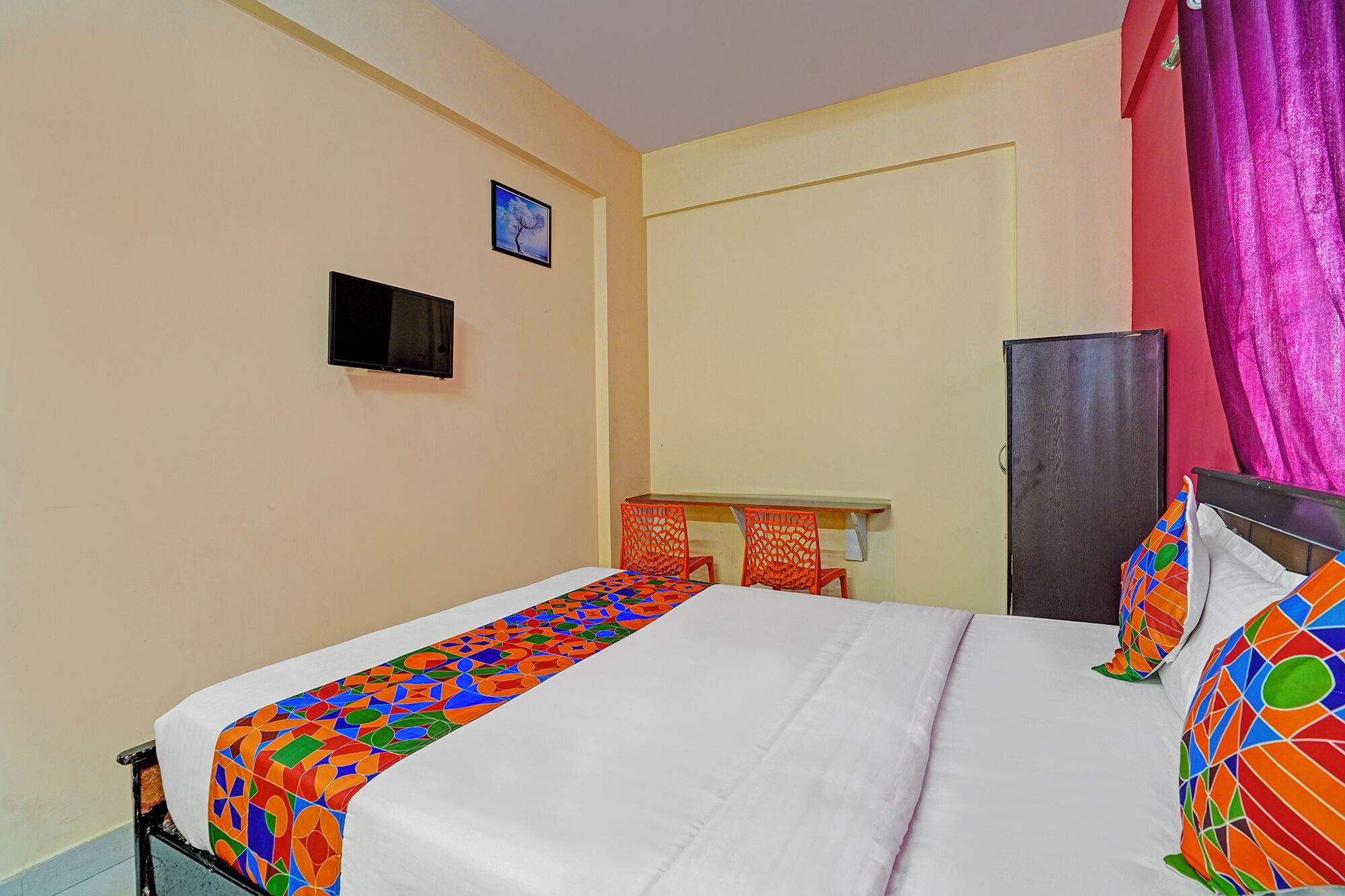 A single bed with a colorful geometric-patterned bedspread, a small desk with a chair, a television mounted on the wall, and a door with a pink curtain.