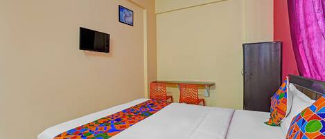 A single bed with a colorful geometric-patterned bedspread, a small desk with a chair, a television mounted on the wall, and a door with a pink curtain.