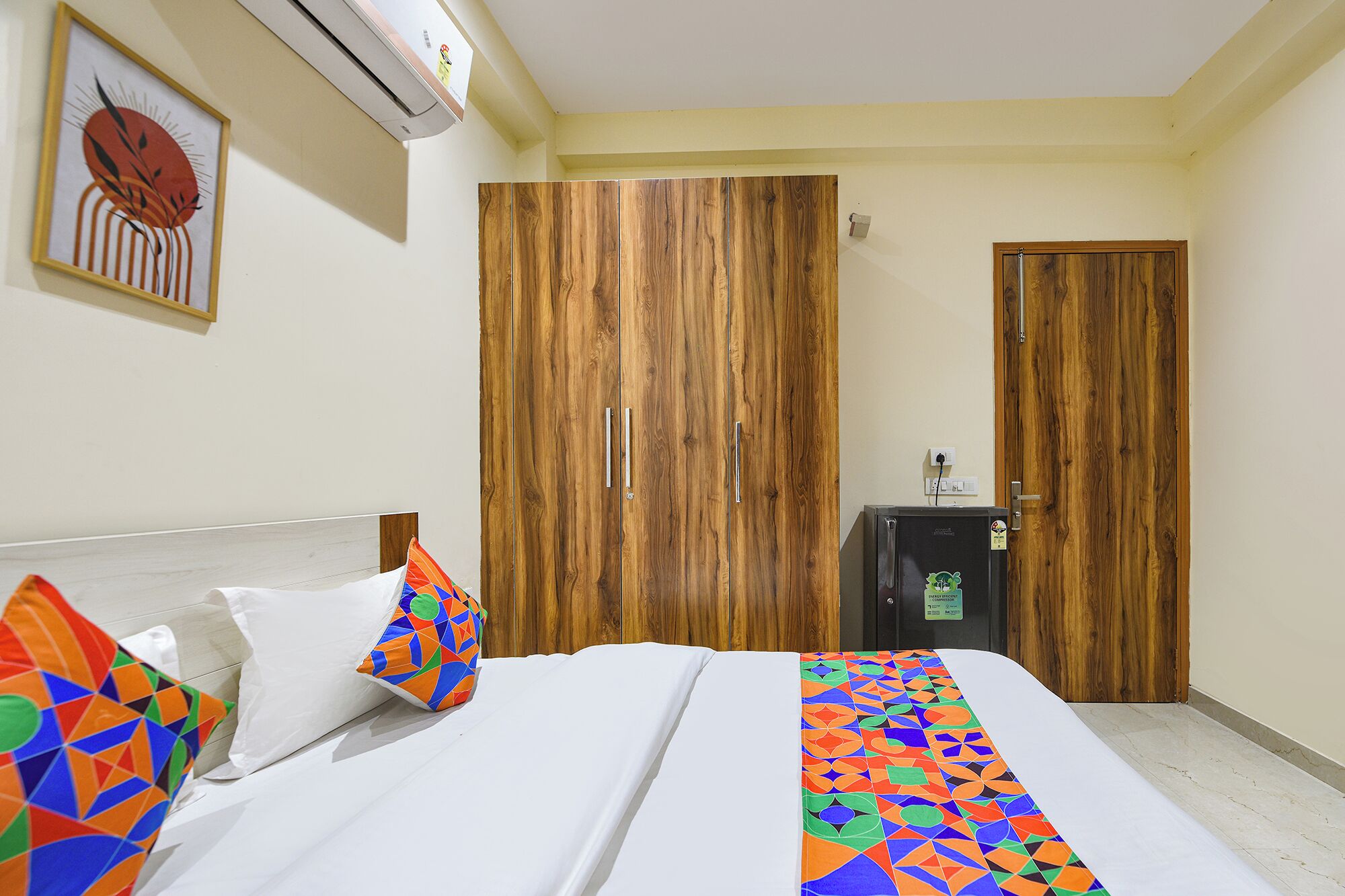 Een hotelkamer met een bed, kleurrijke kussens, een houten garderobe, een bureau en een airconditioning.