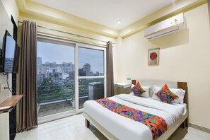 A hotel room with a bed, a TV, a window with curtains, an air conditioner, and a city view. - FabHotel Sunshine I (Gurugram)