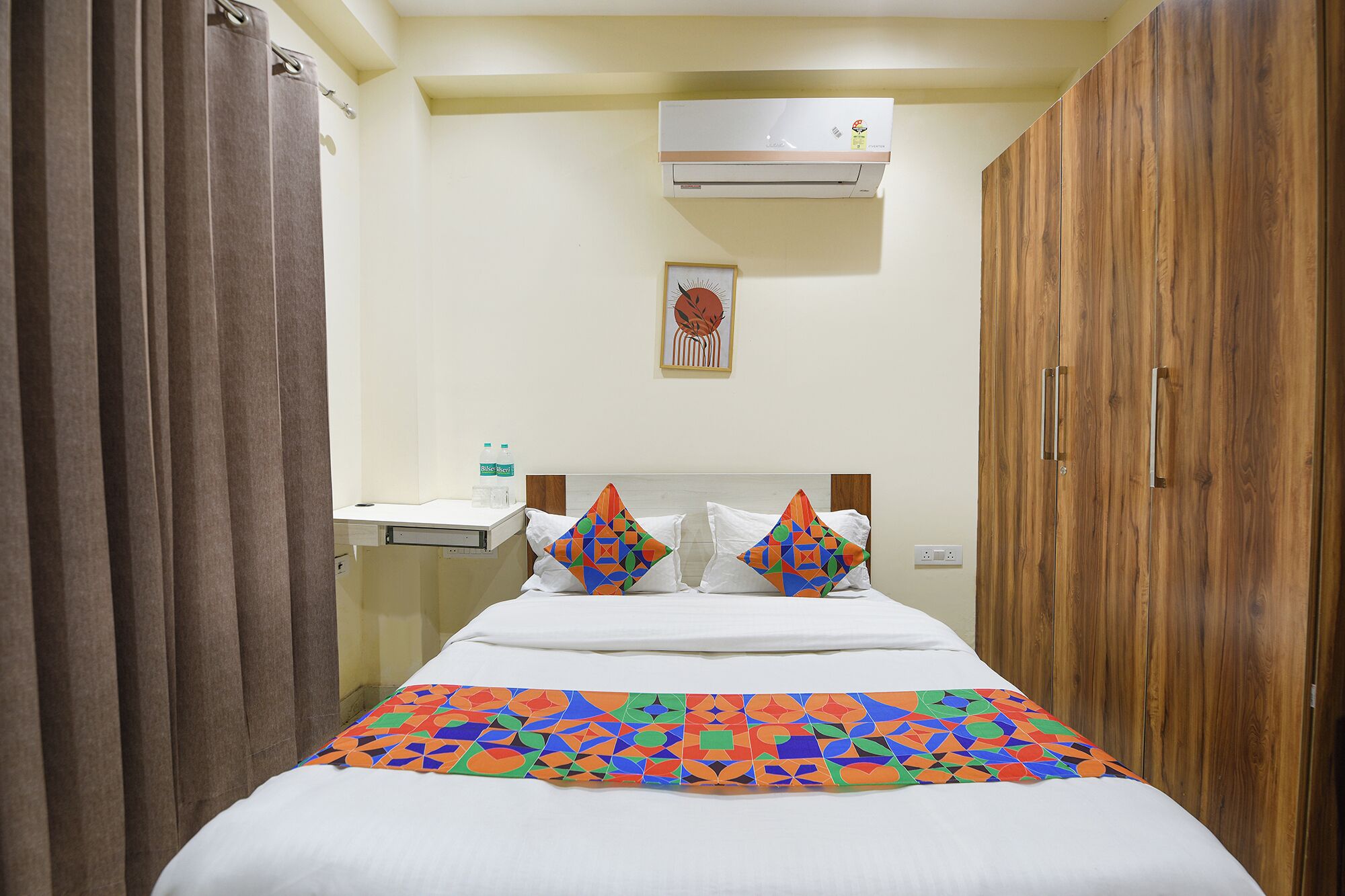 Ein Hotelzimmer mit Bett, Holzschrank, Wand-Klimaanlage, Nachttisch mit einer Wasserflasche und einem gerahmten Bild an der Wand.