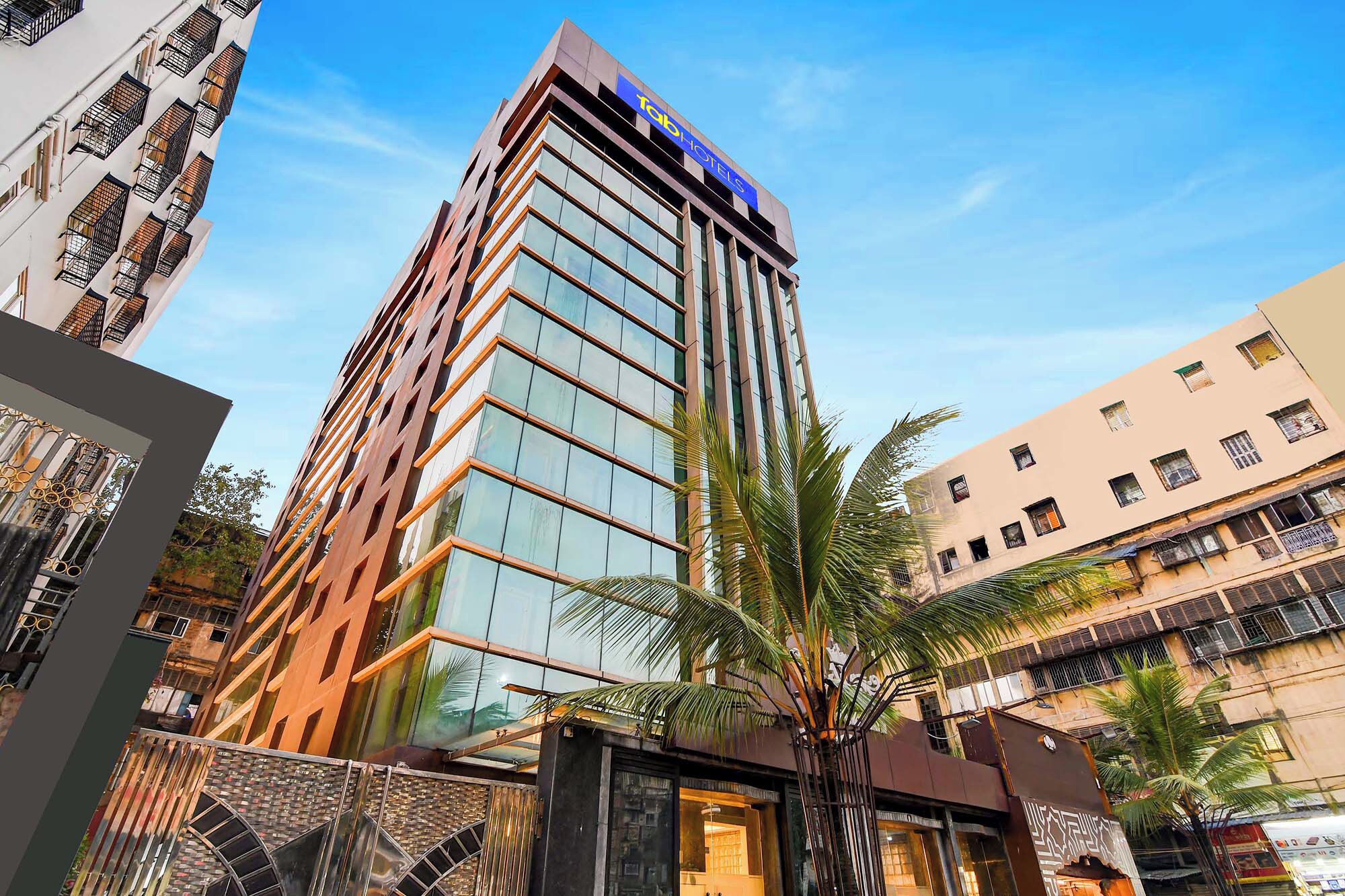 A modern hotel building with a prominent 'Tab Hotels' sign, large windows, and a palm tree in the foreground.