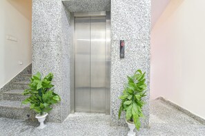 Un ascenseur dans un couloir aux murs recouverts de granit, avec deux plantes en pot et un escalier sur le côté gauche.