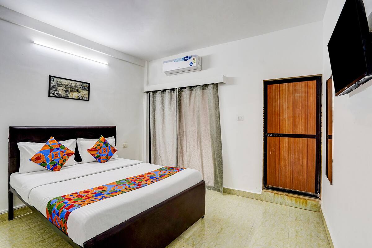 A hotel room with a bed, a flat-screen TV, an air conditioner, a wooden door, and a framed picture on the wall.