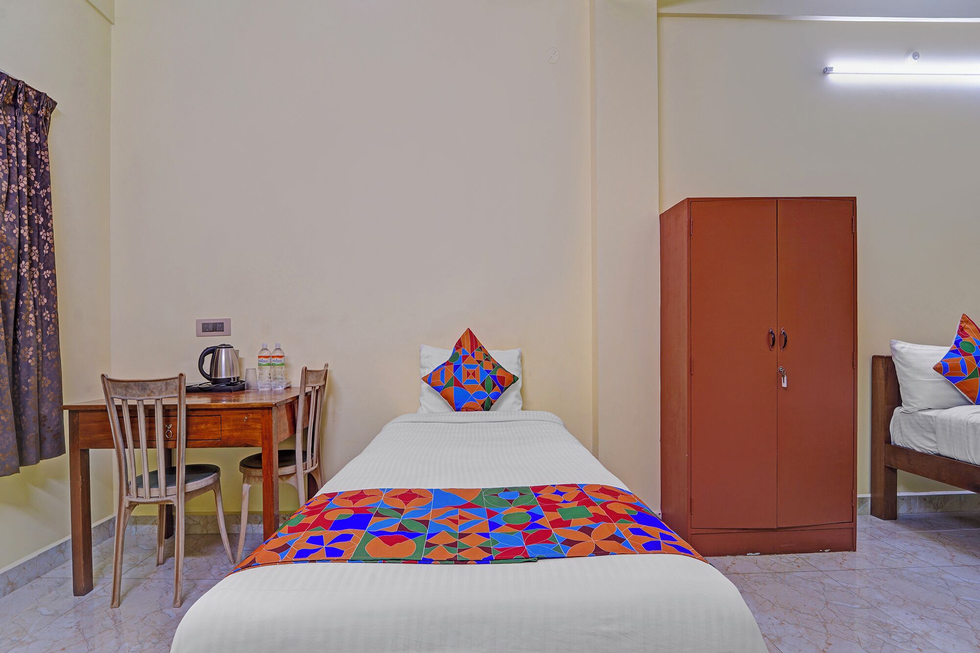 Habitación de hotel con cama, escritorio de madera con tetera, dos botellas de agua, almohada colorida y armario.
