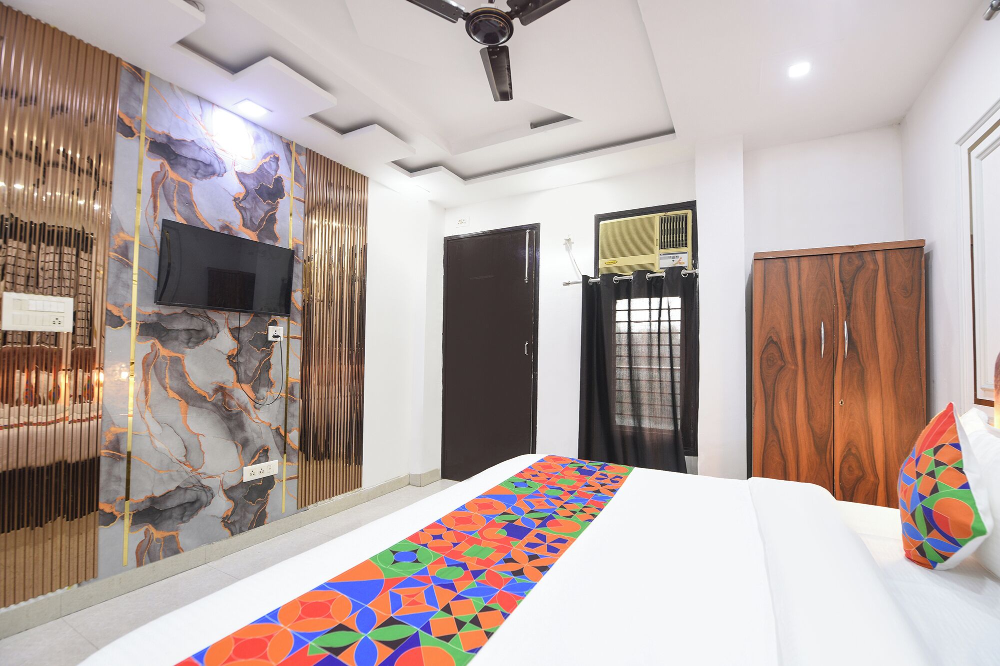 A hotel room with a bed, a TV mounted on the wall, a ceiling fan, and a window with curtains.