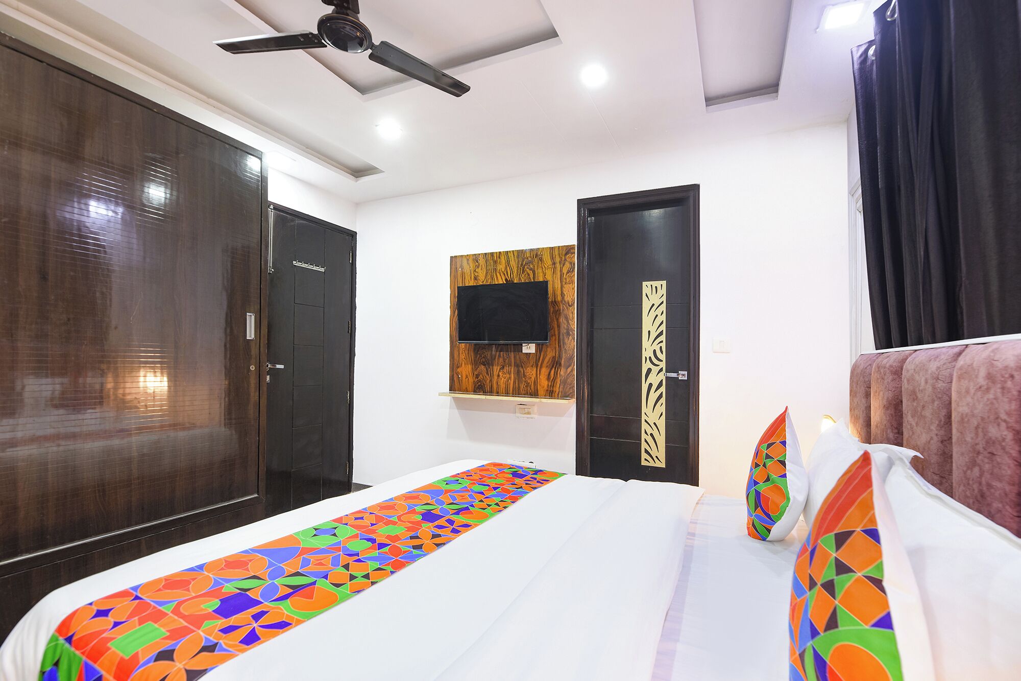 A hotel room with a large bed, a ceiling fan, a TV mounted on the wall, and a door with a decorative panel.