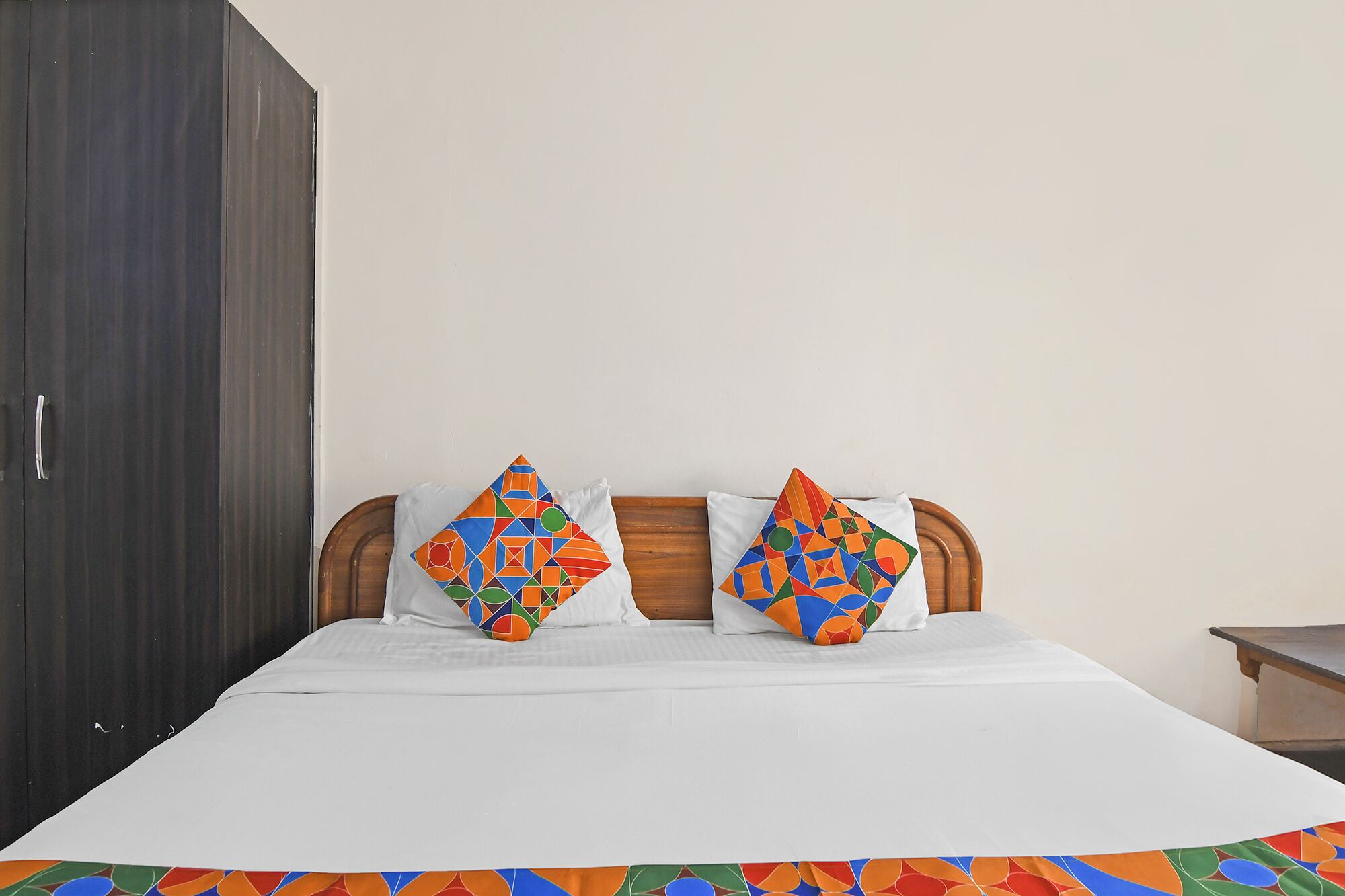 A bed with white bedding, two colorful geometric-patterned pillows, a wooden headboard, and a dark wardrobe.
