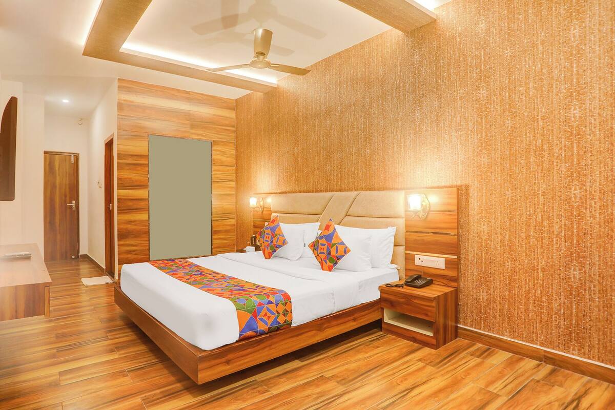 A modern hotel room with a wooden floor, a bed with white bedding and colorful geometric pillows, a nightstand with a phone, and a wall-mounted light fixture.