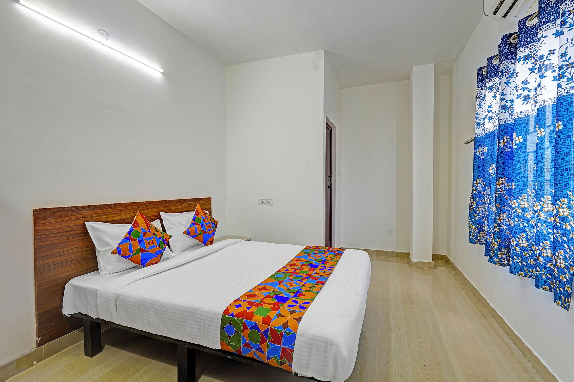 A bedroom with a wooden headboard, a bed with white bedding, colorful geometric pillows, and a blue floral curtain.